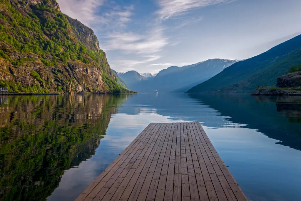 Serene view of Aurland fjord in Norway featuring a tranquil lake bordered by steep mountains, with a wooden dock leading into calm waters under a clear blue sky.