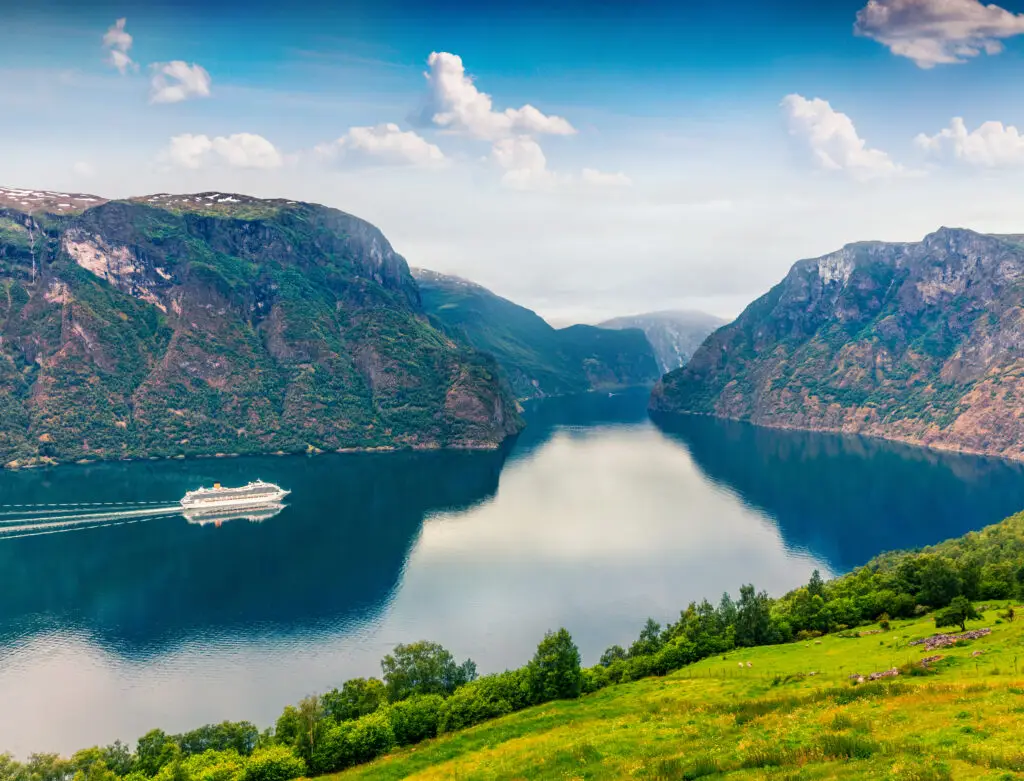 Summer view of Sognefjorden fjord with a cruising ship, surrounded by green covered mountains under a cloudy blue sky, Aurlandsvangen, Norway.
