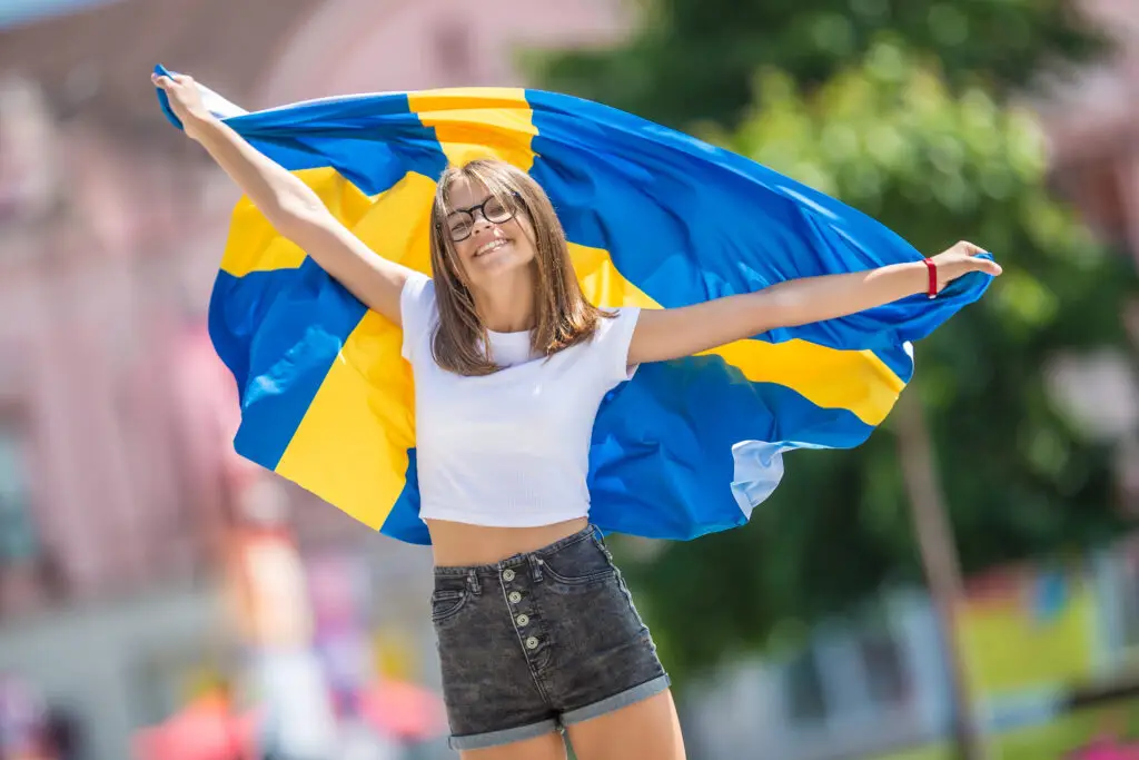 Joyful female tourist celebrating with a large Swedish flag on a sunny street in Sweden, enhancing the festive atmosphere of Scandinavian travel.