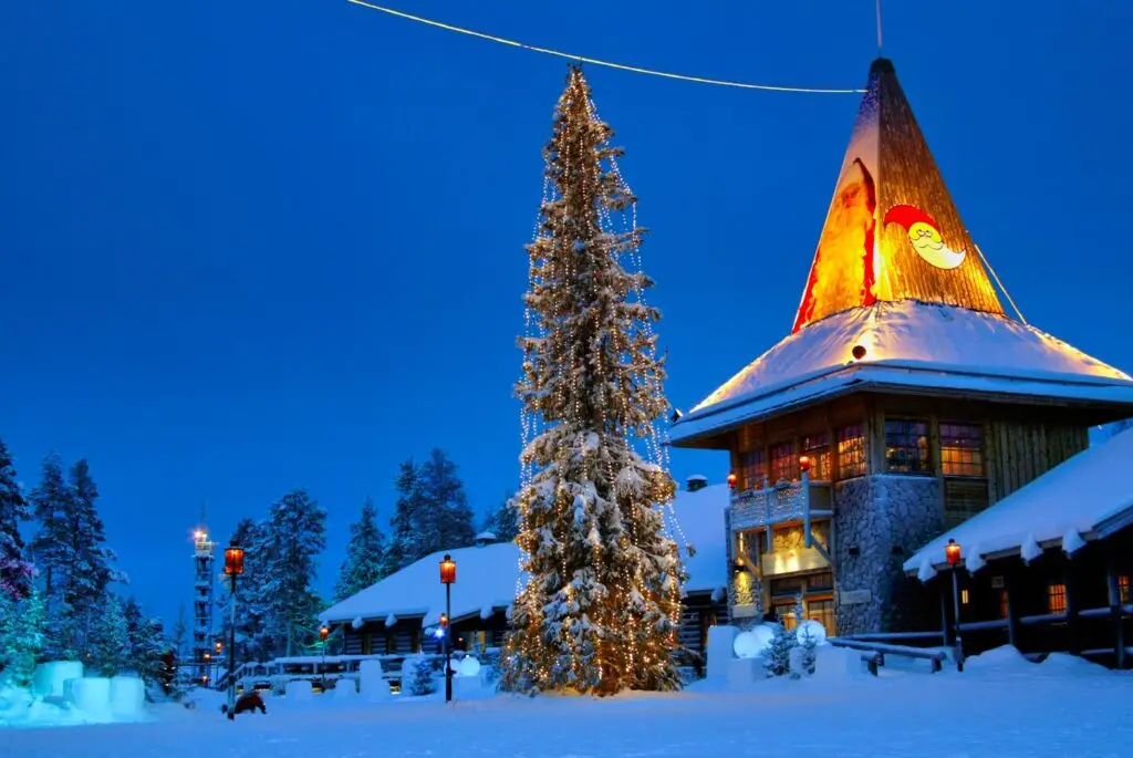 Santa Claus Village in Rovaniemi, Finland, featuring a snow-covered Christmas tree and a warmly lit building with a Santa image at twilight.