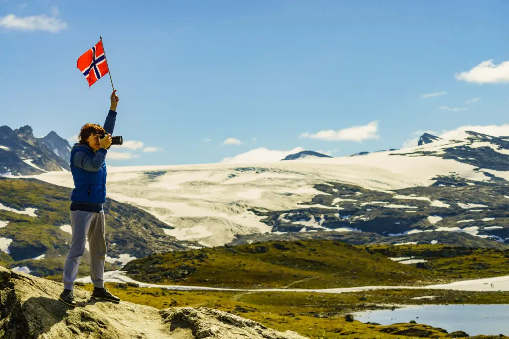 Tourist waving Norwegian flag while taking photographs in the scenic mountains of Norway, showcasing breathtaking views of snow-covered peaks and lush valleys