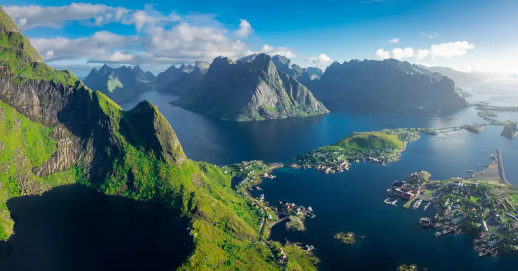 Aerial view of Reinebringen mountain with Lake Reine and the Lofoten Islands village bathed in sunlight, highlighting Norway's breathtaking natural landscapes.
