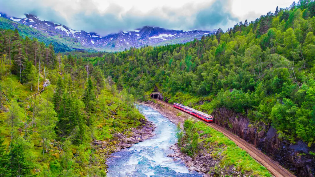 Scenic view of a red train traveling from Oslo to Bergen through lush green mountains and alongside a vibrant blue river in Norway.