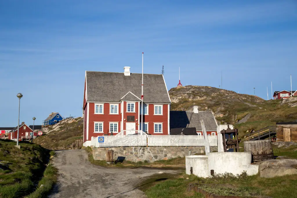 Knud Rasmussen Museum in Ilulissat, Greenland, featuring red colonial architecture with a lush, rocky landscape under a clear blue sky.
