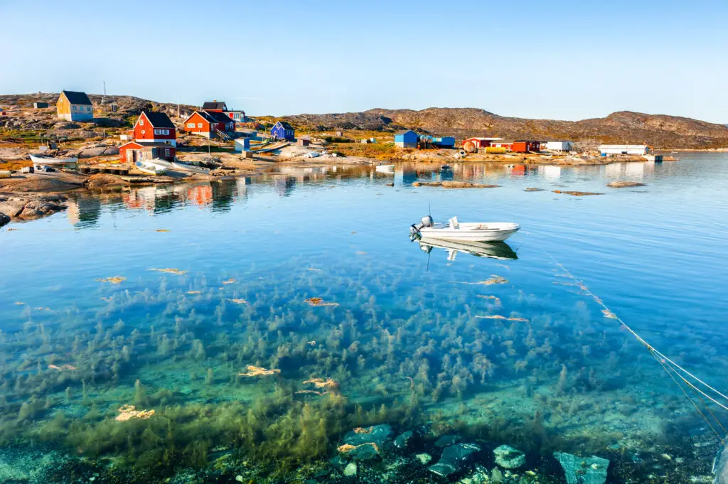 Colorful houses of Oqaatsut village on rocky Greenlandic coast with clear blue waters and a moored boat, reflecting summer in Greenland.