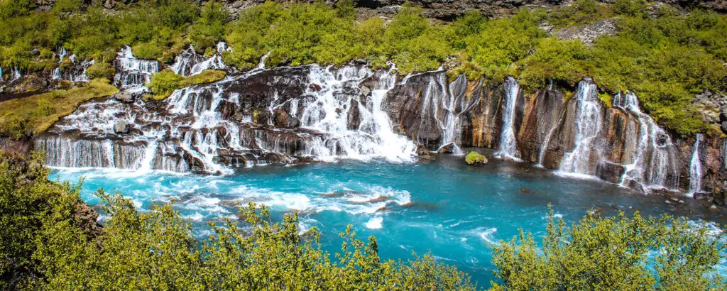 Stunning view of Hraunfossar waterfalls in Iceland with cascading water over mossy rocks and vibrant turquoise river surrounded by green foliage.