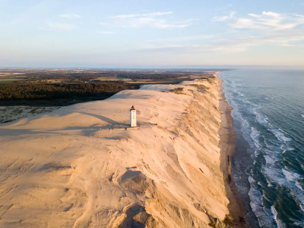 Aerial view of Rubjerg Knude Lighthouse surrounded by sand dunes on Denmark's North Sea coast, a scenic landmark popular with tourists.