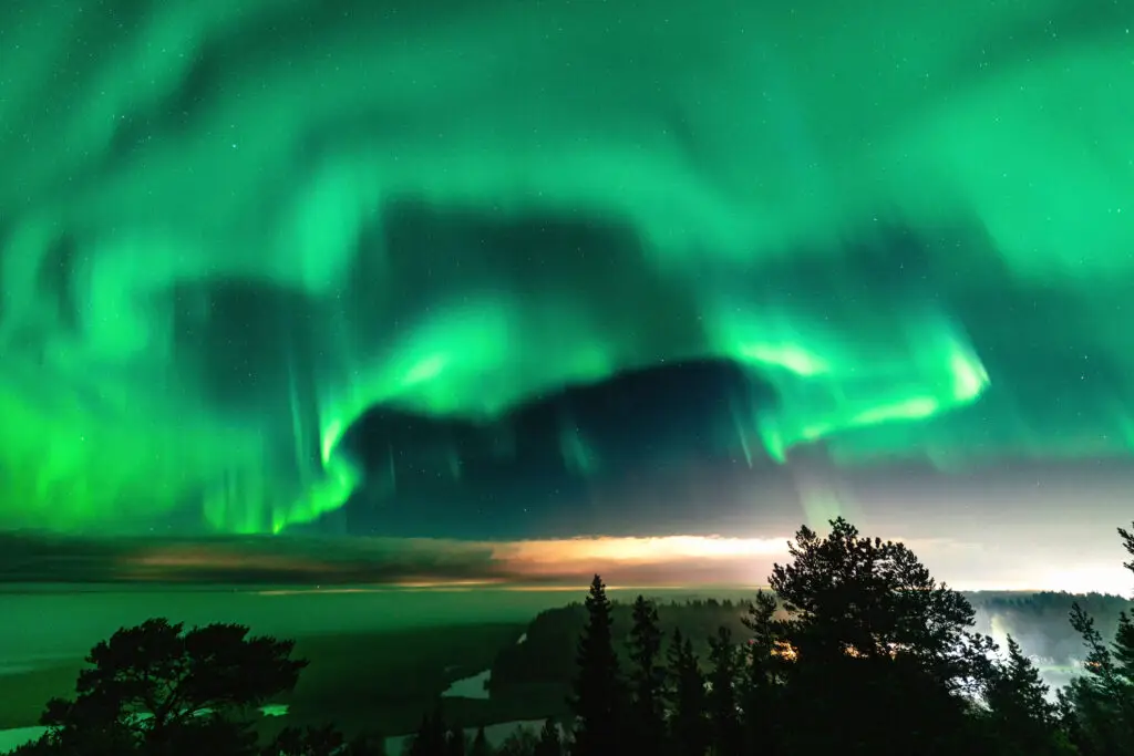 Brilliant green Aurora Borealis over a foggy forest and mountainous landscape with light rays from a village in Northern Sweden, perfect for a unique Scandinavian tour experience.