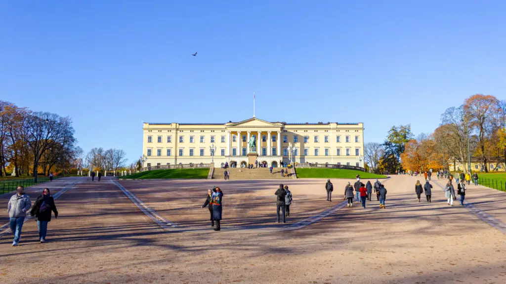 Tourists walking towards the Royal Palace in Oslo Norway on a sunny day, showcasing the majestic facade and autumn trees.