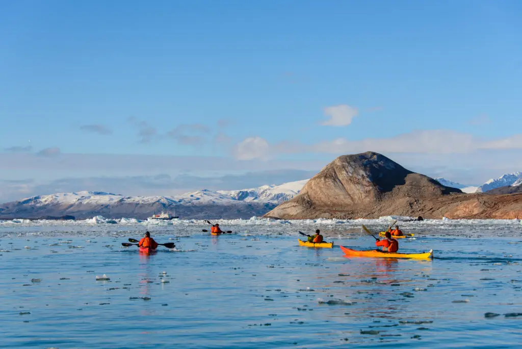 Tourists kayaking among icebergs in Greenland with sunny summer skies and mountainous backdrop, showcasing Arctic adventure tourism.