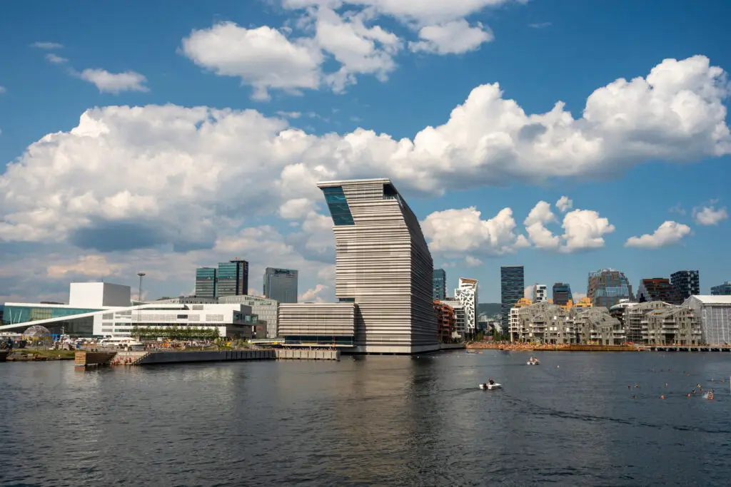 Summer day at Oslo's waterfront showing the modern architecture of Munch Museum, surrounded by other buildings with a serene sea in the foreground and a blue sky with fluffy clouds above.