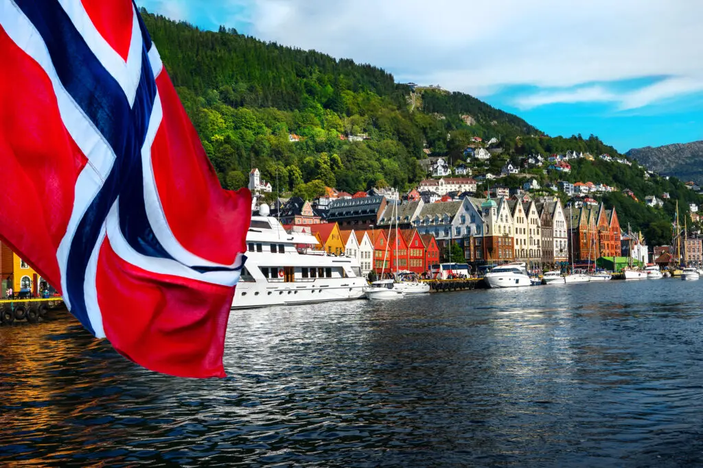 Norwegian flag waving in front of the scenic Bryggen waterfront with traditional colorful houses and boats in Bergen, Norway
