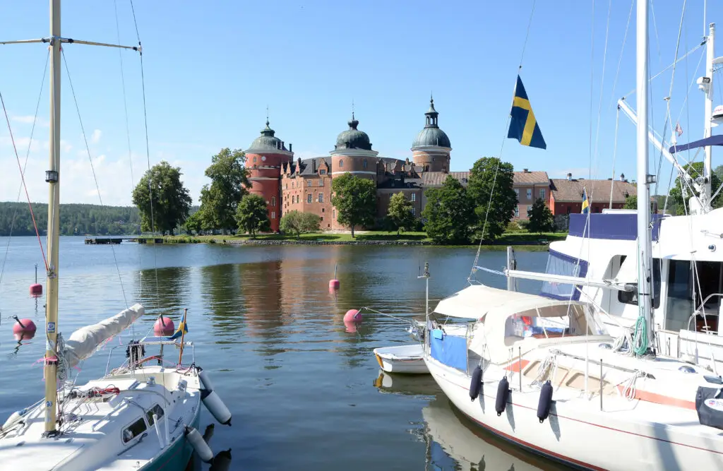 Scenic view of Gripsholm Castle in Mariefred, Sweden, with boats moored in lake foreground and Swedish flag fluttering on a sunny day.