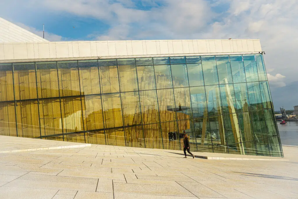 Tourist walking by the modern architecture of Oslo Opera House in Norway, reflecting cityscape in its glass facade.
