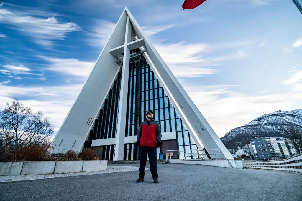 Young man with beard in red jacket standing in front of the iconic Arctic Cathedral in Tromso, Norway, with scenic snowy mountains in the background.