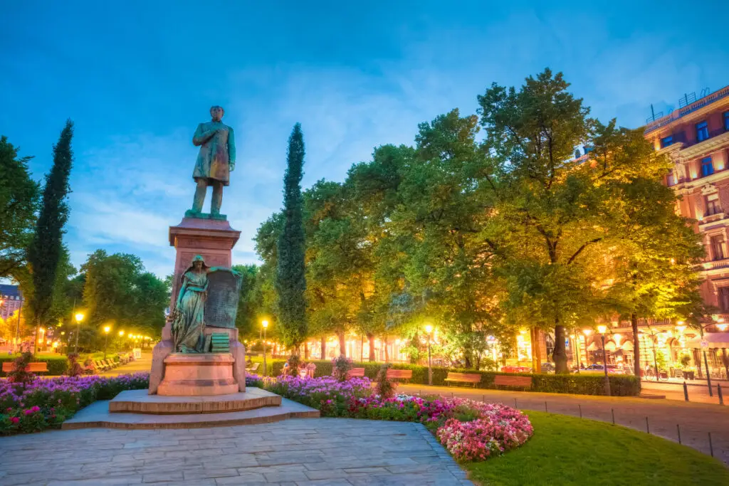 Statue of Johan Ludvig Runeberg in Esplanade Park, Helsinki surrounded by illuminated trees and flower beds at twilight.