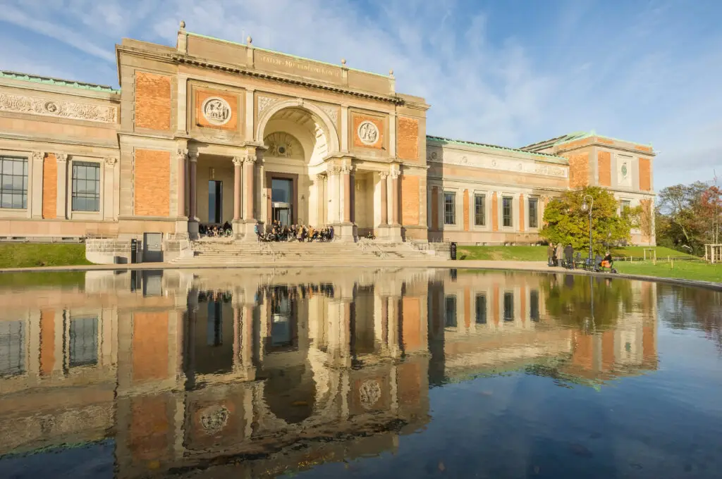 Exterior view of the National Gallery of Denmark in Copenhagen, showing its reflection on a serene pond with visitors enjoying the sunny day