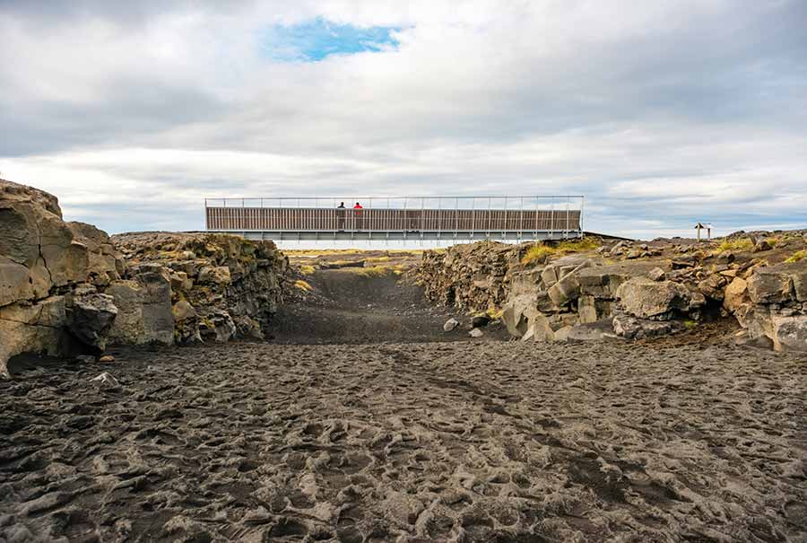 Tourists walking on the Bridge Between Continents over a rugged rift valley in Iceland, showcasing the geological marvel where the Eurasian and North American tectonic plates meet.