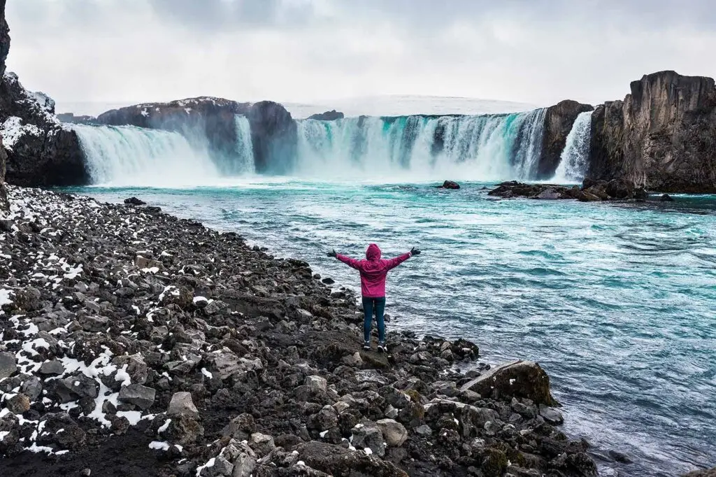 Tourist with arms outstretched at the majestic Godafoss Waterfall in Iceland, experiencing the raw natural beauty on a custom Scandinavia tour.