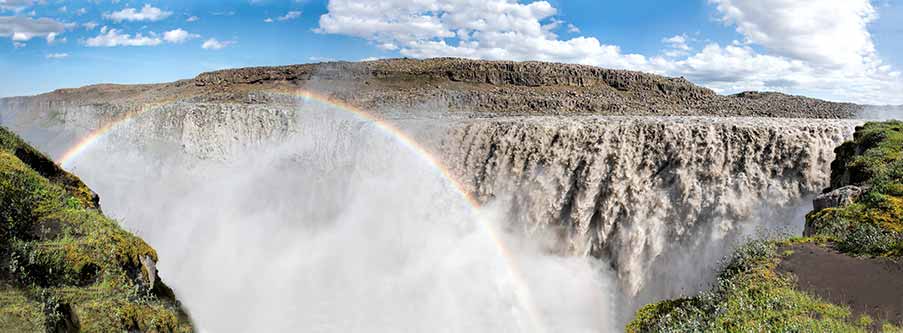 Panoramic view of Dettifoss Falls in Iceland, featuring a powerful waterfall with rising mist and a vivid rainbow over rocky terrain.