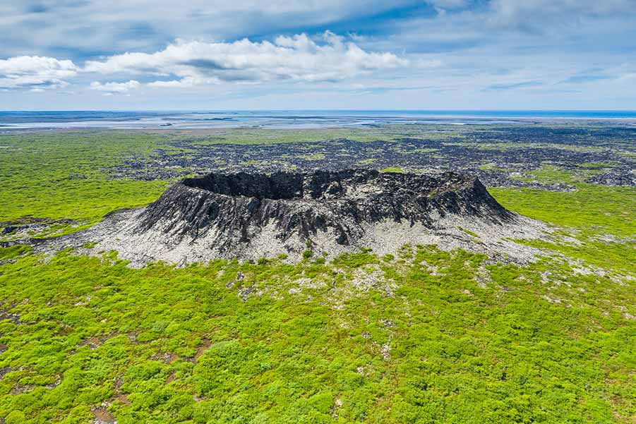 Aerial view of Eldborg Crater in Iceland, showcasing the lush green landscape surrounding the distinct black volcanic crater under a vast blue sky.