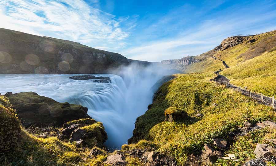 Breathtaking view of Gullfoss Waterfall in Iceland, showcasing the powerful cascades of water and lush greenery in a sunny, scenic setting.