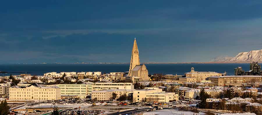 Panoramic view of Hallgrímskirkja church dominating the skyline of Reykjavik, Iceland, with snowy mountains in the background and a coastal view, perfect for cultural tourism enthusiasts.