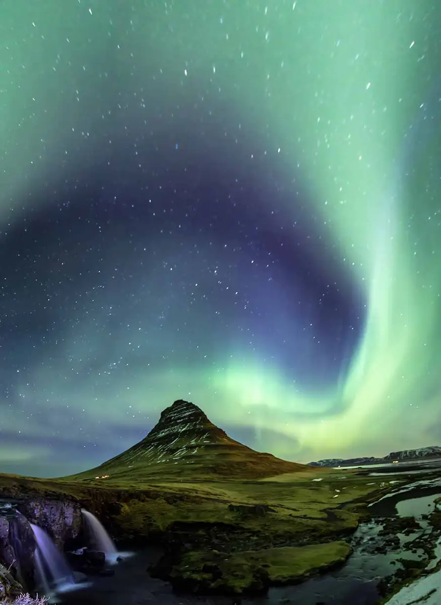 Stunning view of the Northern Lights swirling above Kirkjufell mountain in Iceland, with a starry sky and flowing waterfall in the foreground.