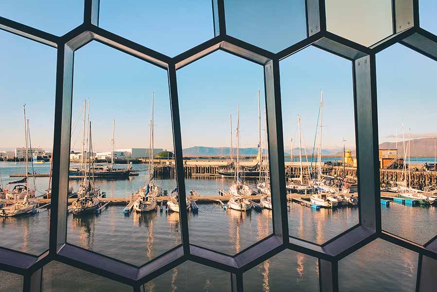 Panoramic sea view of Reykjavik harbor through the geometric glass facade of Harpa Concert Hall in Iceland, showcasing moored sailboats and distant mountains.