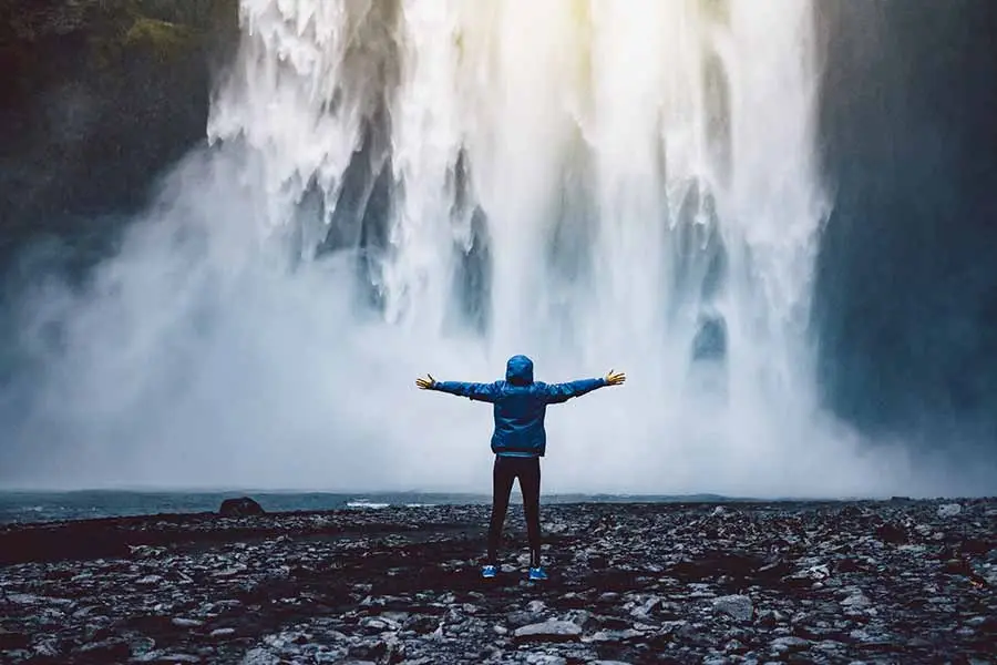 Tourist with outstretched arms facing the majestic Skogafoss waterfall in Iceland, showcasing the natural beauty and scale of the attraction.