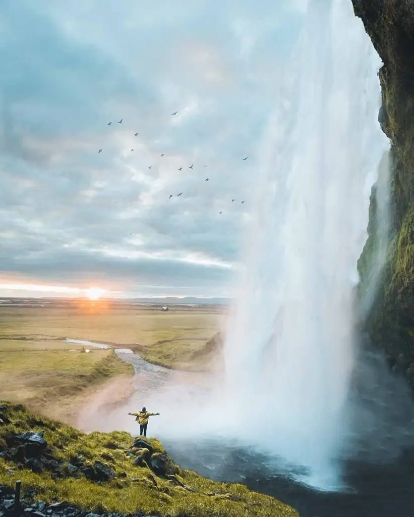 Tourist enjoying a breathtaking sunset at a majestic waterfall in Iceland, with birds flying overhead and a serene river landscape in the background.