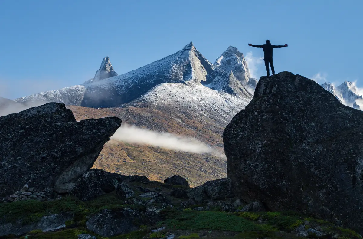 Person standing with outstretched arms on a rock at Ulamertorsuaq Mountain, overlooking the rugged snow-capped peaks and foggy valleys of Tasermiut Fjord in South Greenland.