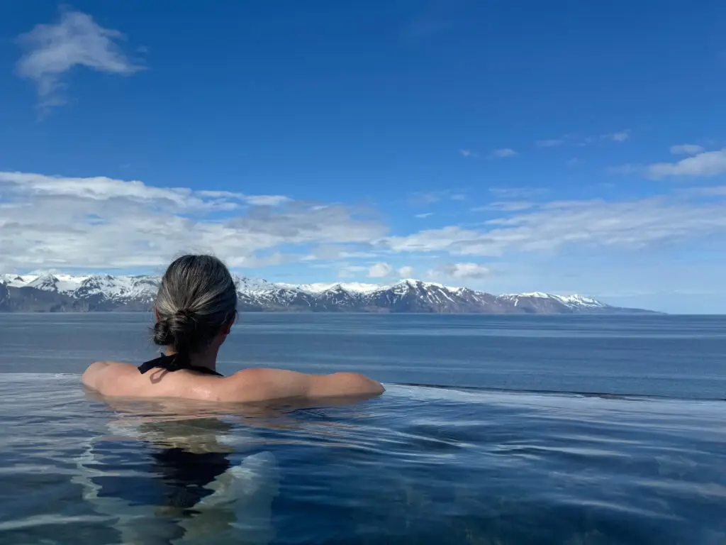 Woman relaxing in a geothermal hot pool overlooking a serene bay and snow-capped mountains in Scandinavia, experiencing the natural beauty and tranquility of the region.