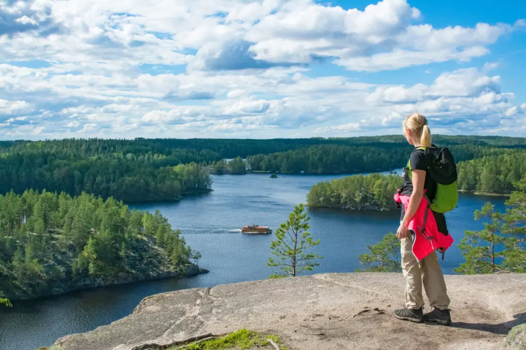 Woman hiking in Finland, viewing a scenic landscape from a high cliff in a National Park, showcasing lush forests and a serene lake.
