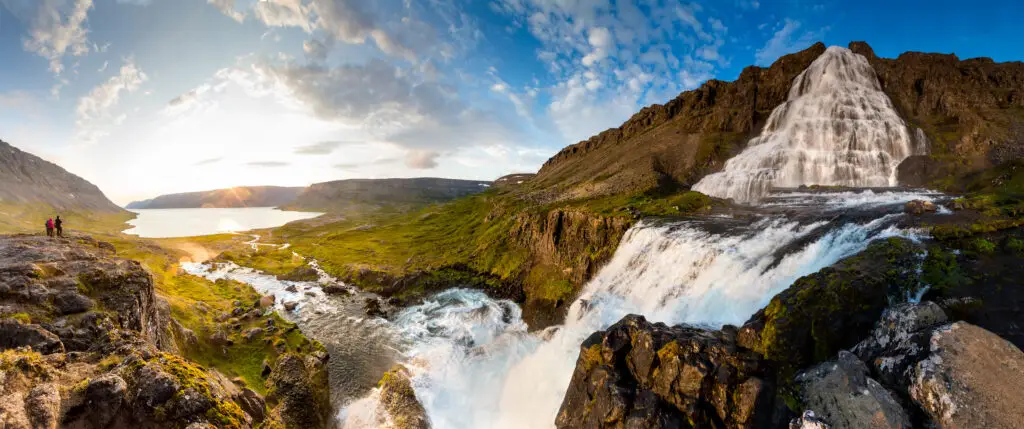 Panoramic view of the majestic Dynjandi waterfall in Iceland, featuring cascades flowing over lush green terrain under a clear blue sky.