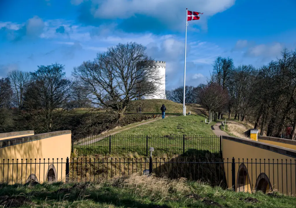 Historic Fredericia fortification in Denmark featuring lush green landscape, a prominent white tower, and the Danish flag fluttering against a blue sky.