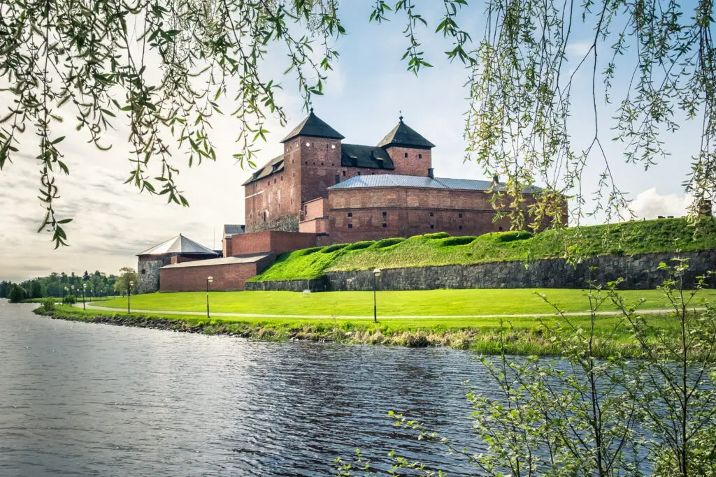 Medieval castle in Hameenlinna, Finland, standing majestically by the water surrounded by greenery and framed by willow branches in spring.