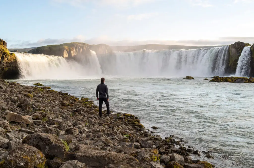 Man observing the majestic Godafoss waterfall in Iceland, surrounded by rugged terrain and cascading waters under a clear sky.