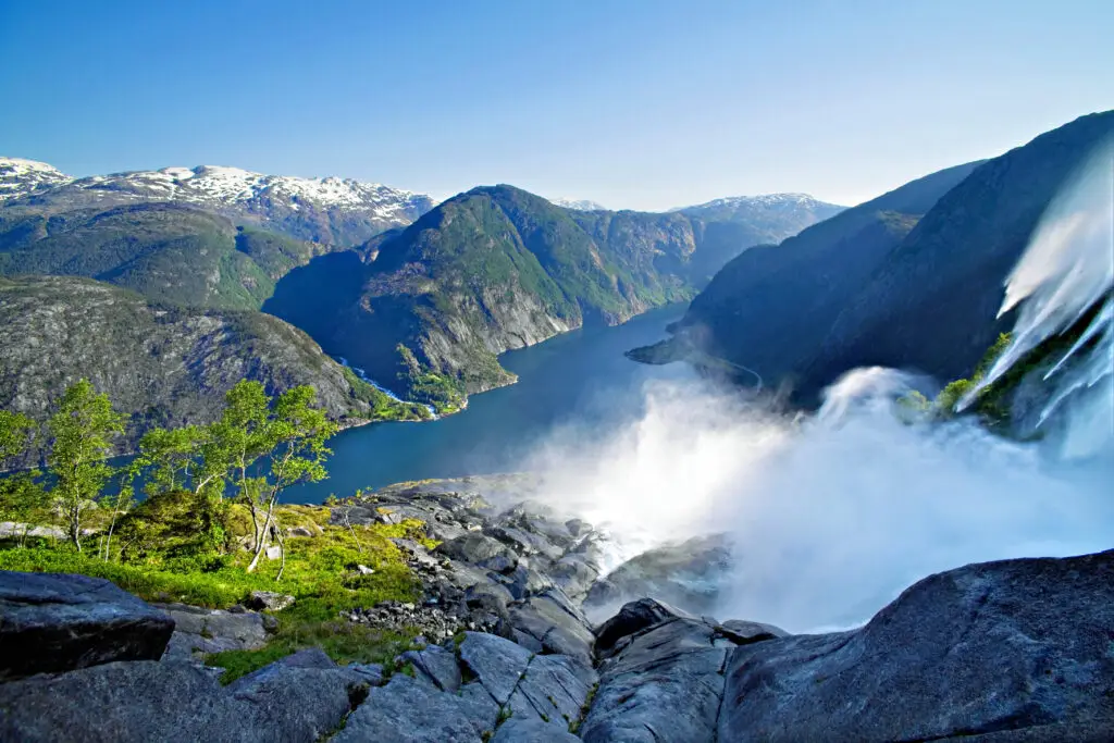 Breathtaking view of Langfoss Waterfall cascading into Akrafjord with lush greenery and snow-capped mountains in the background, Norway.
