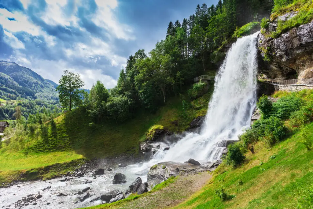 Breathtaking view of a powerful waterfall cascading down jagged rocks into a river in rural Norway, surrounded by lush greenery and scenic mountains, perfect for nature lovers and adventure tourism.