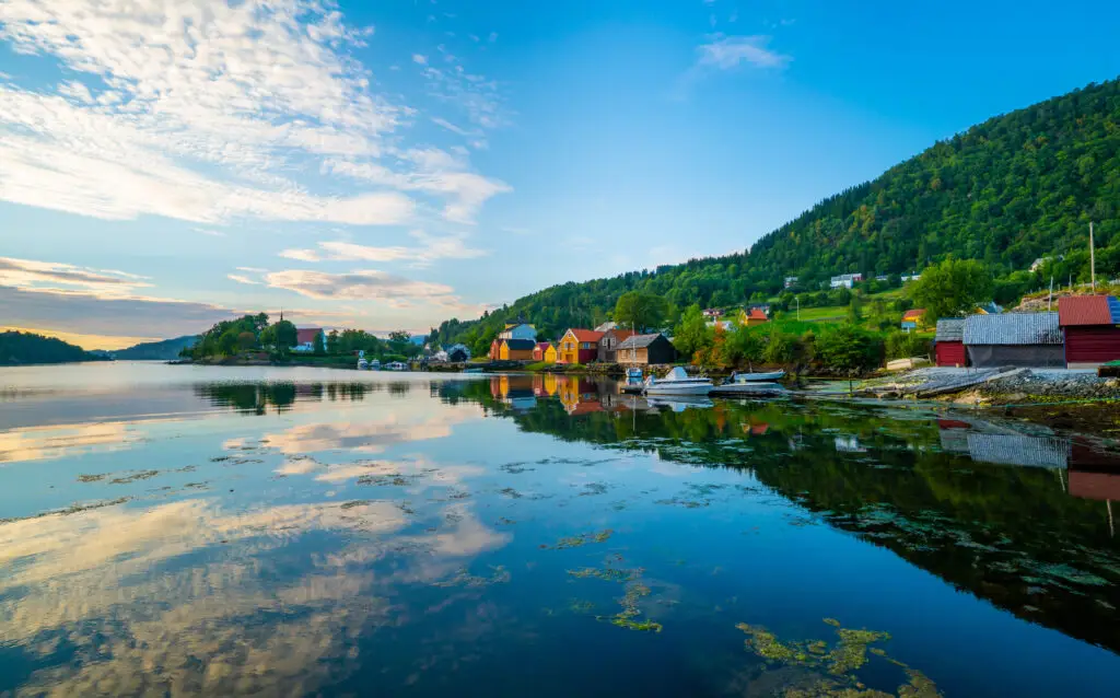 Sundown view of colorful houses reflecting in the calm blue waters of Hosaner harbor on Osterøy Island, Norway, illustrating a scenic Nordic landscape.