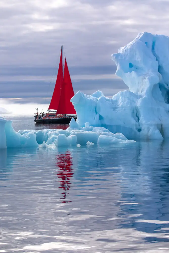 Red sailboat sailing beside a towering iceberg in Disko Bay, Ilulissat, Greenland, showcasing Arctic adventure tourism.