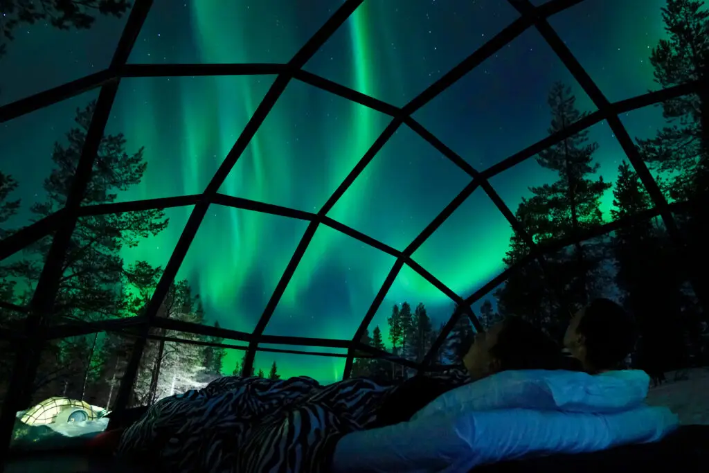 Young Caucasian couple enjoying a view of the Northern Lights from inside a glass igloo in the Scandinavian wilderness, perfect for romantic getaways and unique travel experiences.