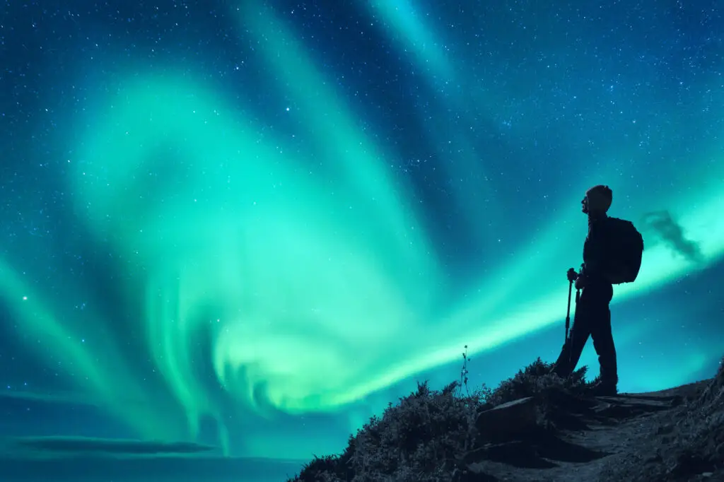 Silhouette of a woman with a backpack observing the stunning Aurora Borealis in the starry night sky, perfect for a trekking tour in Scandinavia.
