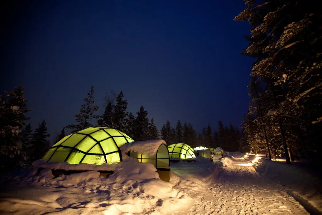 Snow-covered glass igloos illuminated under night sky in Scandinavian winter landscape, ideal for unique tourism and accommodation.