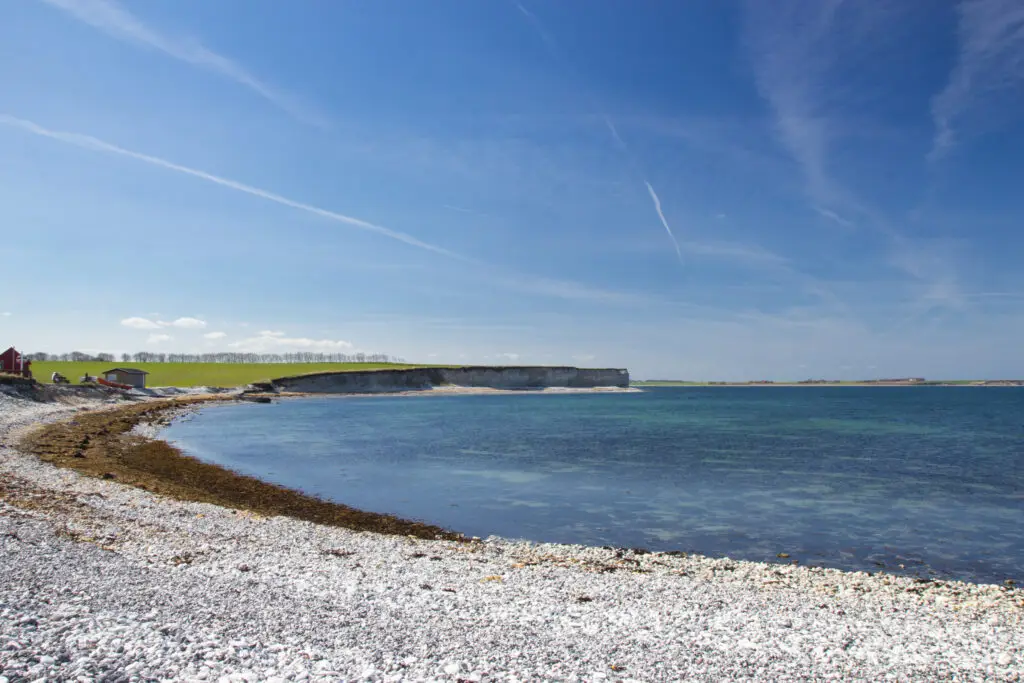 Scenic view of Sangstrup Klint, the white cliffs on Djursland coast, Denmark, showcasing clear blue waters and a pebbled beach under a sunny sky.