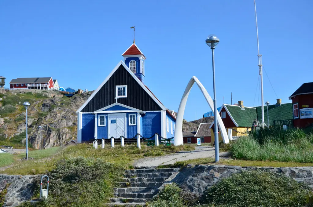 Entrance to the open-air museum in Sisimiut, Greenland featuring traditional colorful wooden buildings and a unique arch structure under a clear blue sky.