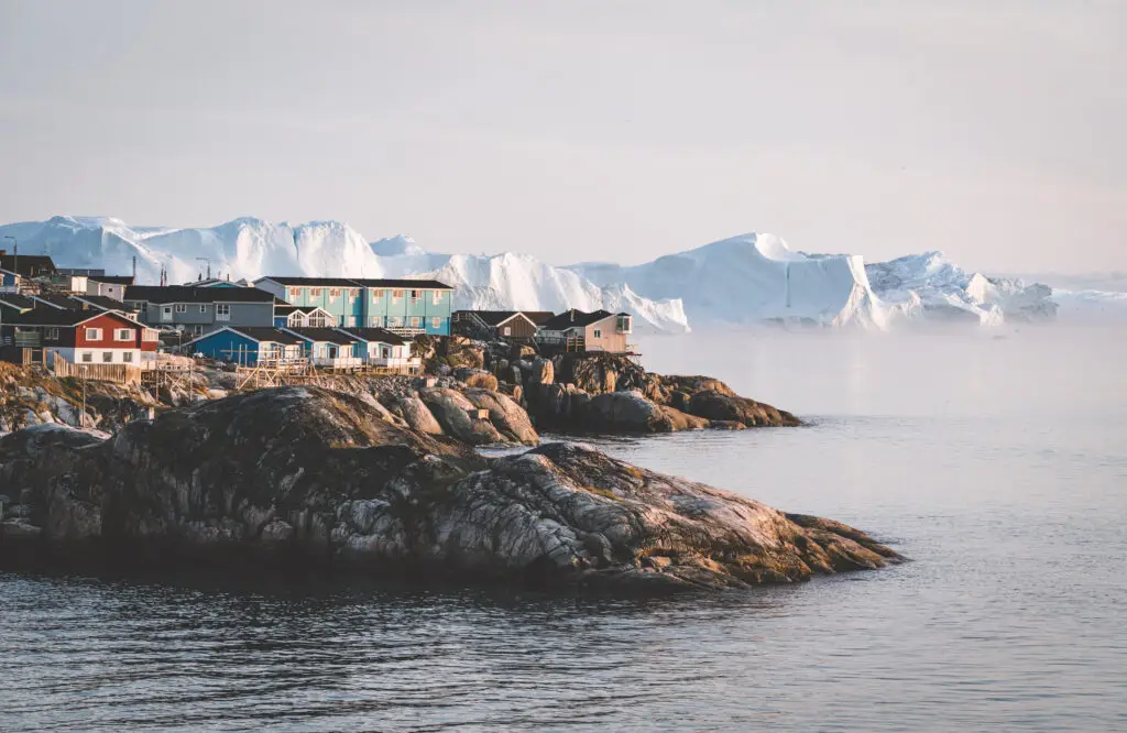 Aerial view of colorful houses in Ilulissat, Greenland, with icebergs and foggy orange-pink sunrise sunset in summer.