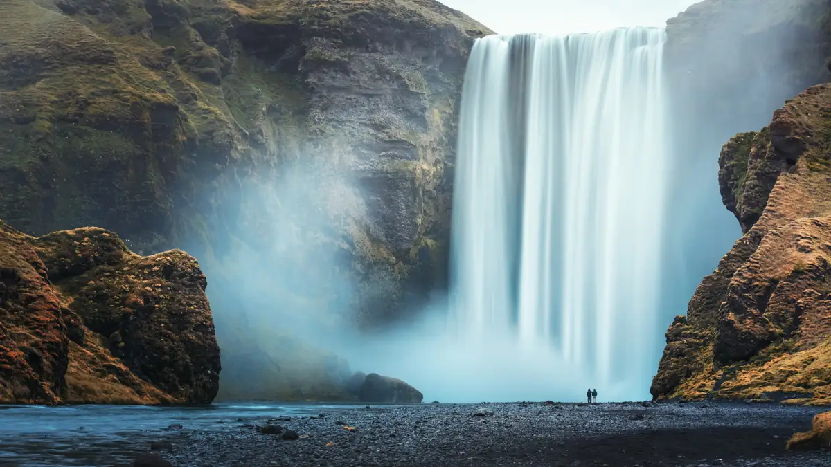 Tourist couple near the majestic Skogafoss waterfall in Iceland, showcasing the powerful cascade and misty ambiance of the scenic landscape.