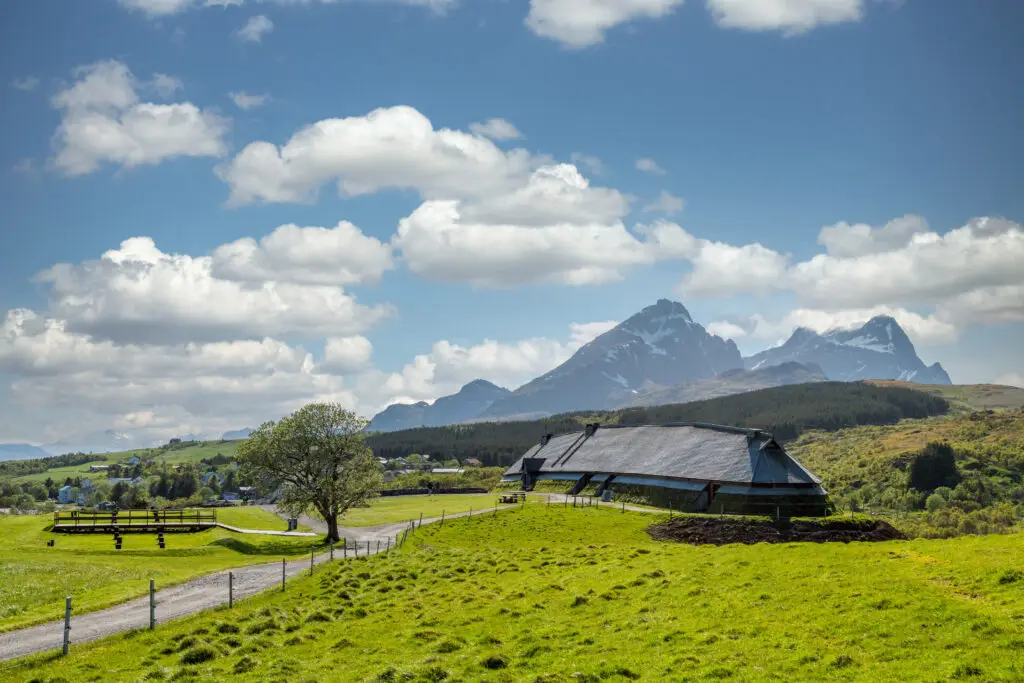 Lofotr Viking Museum Longhouse with scenic mountain backdrop and lush green fields under a blue sky with white clouds, epitomizing Viking era architecture and history in Scandinavia.